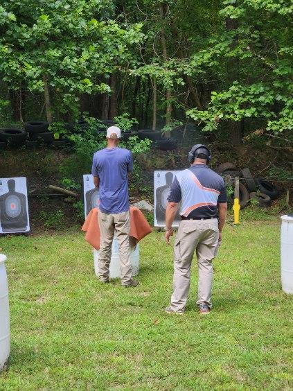 A young African-American male in a blue shirt and khakis points a pistol at a target while an older Filipino man in a two-tone blue shirt and khakis watches on.